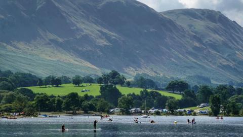 Ullswater in the Lake District, with the high fells on the horizon. There are several people paddleboarding and swimming in the lake. It is a summer's day, where there is bright green grass and several trees.