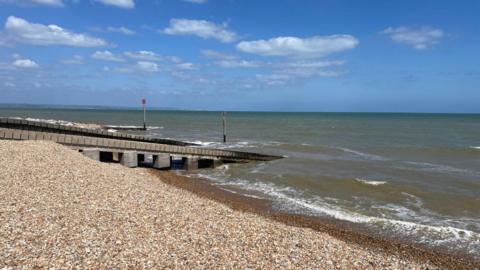 A pebble beach. The seawater is dark and muddy. The sky is bright blue. 