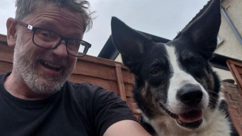 Chris Durham, a man with a grey beard, glasses and a dog are stood outside next to a brown wooden garden fence