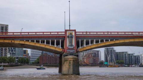 A red and yellow bridge with a statue positioned on one of the supports. The background shows a skyline of high rise buildings. 