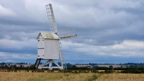 A white windmill in the foreground with three sails with a hill in the background spotted with trees underneath a bright blue sky full of clouds 