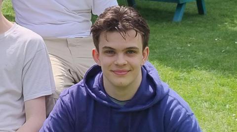 Max Prince, a young man with dark spiky hair wearing a purple hoodie, sitting on grass and smiling 