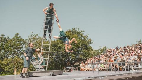 A group of six performers hold up a ladder as one stands at the top and another dangles from it. They are watched by an audience sitting on a stand under a blue sky.