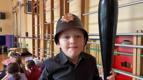 A boy is dressed up like a policeman. He is holding an inflatable truncheon and a plastic police helmet. He is standing in a school gymnasium and other children are seated on the floor behind him