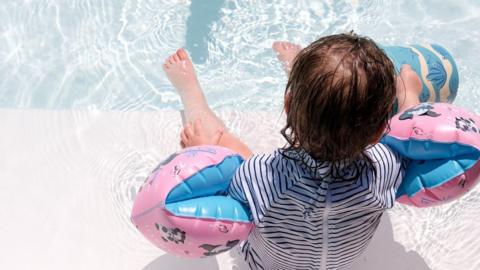 A toddler with dark, wet hair, who is wearing a black and white striped swimsuit and pink and blue armbands, sits on the edge of a swimming pool with their feet in the water