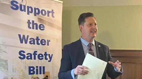 Mp Lee Pitcher standing in a room with wood-panelled walls. He is wearing a dark suit and his body is angled to the right, with his eyes looking in the same direction. He is a document in his right hand and gesturing with the other while speaking. On the left, a tall banner reads “Support the Water Safety Bill".