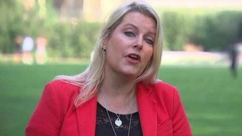 A picture of Mims Davies being interviewed on College Green in Westminster in bright sunshine wearing a red jacket in front of a blurred background of grass and trees