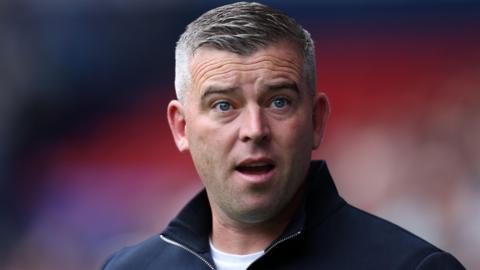 Bolton boss Steven Schumacher, wearing a navy blue half-zip fleece and white t-shirt, stands with his mouth open during a pre-season friendly against Everton