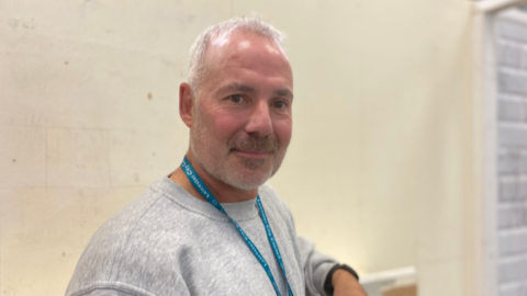 Lee Beeching stands in front of a cream brick wall in a warehouse. He has short grey hair and a short grey beard. He is wearing a grey sweater and has a blue Leicester City Council lanyard.