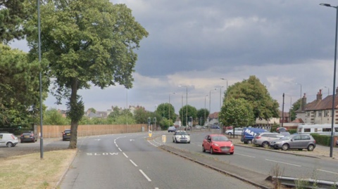 A main road with cars driving along, to the left is a grass verge with a tree, and further in the distance a long stretch of fence, on the right are rows of houses with cars parked in front of them