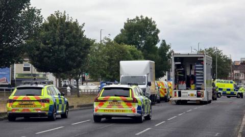 A number of police vehicles close off a dual carriageway in Merseyside. The road has trees lined down the middle. There is a white police van in the middle of the road with its back doors open showing a lot of equipment inside and police tape in front of it where a couple of officers in hi-vis uniform stand.
