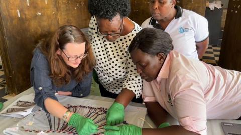 Four women are gathered around a table all looking down at glass headband resting on the table on a white cloth. They at Uganda Museum in June 2024. Three of the women, all wearing green plastic gloves, are pointing at or moving the headband. The fourth stands slightly behind them, scrutinising their movements