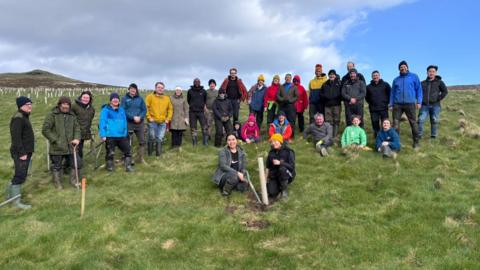 A group of volunteers who plant trees stand for a photo around the final stump of tree they planted.