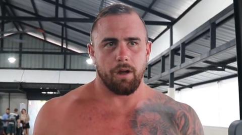 A topless Ben Ransome in the gym. He has short dark hair and a beard, as well as a tattoo on his chest.