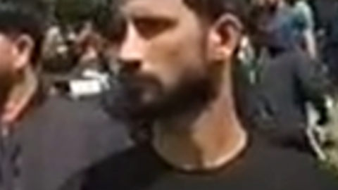 A close-up of a man with black hair and a black beard, with his head turned to the side. He is wearing a  black t-shirt and numerous people are in the background