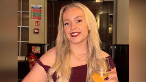 Jessica Thorpe smiling at the camera. She had long blond hair and is wearing a burgundy dress, with matching burgundy painted nails. She is holding a wine glass.