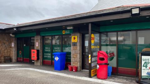 A picture of a service station. There is a red bin and a blue bin in front of the building. There are large windows with green stickers. The sky is grey. 
