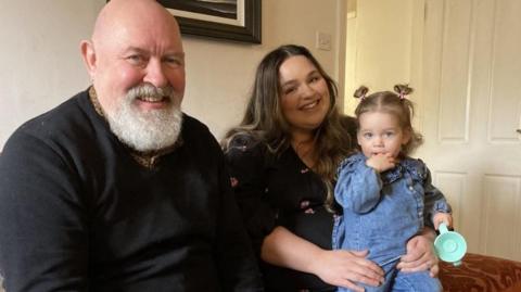 A man, with a bald head and a grey beard, sits on a sofa next to a woman with long, dark hair who has a small female child - dressed in denim - on her knee