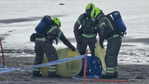 Firefighters wearing protective gear examine a blue drum on a beach. They are holding yellow drums next to the blue drum.