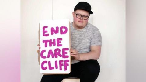 Oscar sits on a stool. He is wearing a black cap and glasses. He is holding a sign that says "end the care cliff"