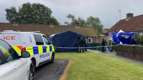 Two blue police tents and a cordon are set up outside bungalows. A police van is parked beside the scene.