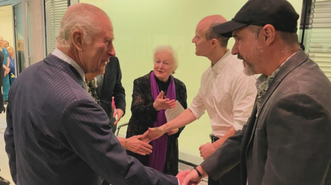 On the left is King Chales III wearing a navy and white pinstripe suit. He has grey hair, and is holding his hand out shaking it with Steve Young. Steve is wearing a grey suit jacket and is wearing a black baseball hat. 