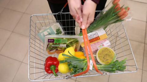 A close-up of a supermarket basket with assorted food items, including lemons, a pepper and spring onions. The hands of the shopper holding the basket can be seen at the top of the picture.