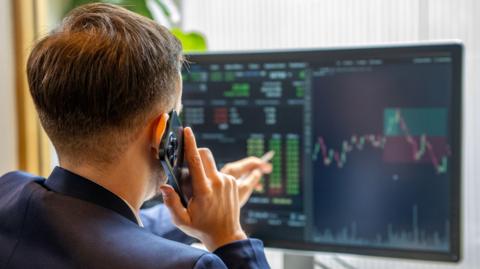 A man holds a mobile phone to his ear while pointing at a large computer screen that is displaying charts and numbers.