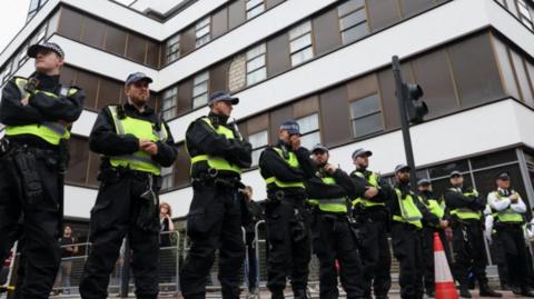 A row of police lined up on a street outside of a tall building 