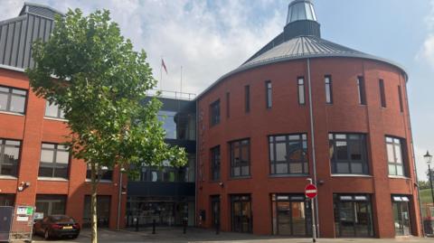 A council building made out of red brick. The building is curved and has grey-framed windows. There is a tree outside and a no entry sign. A car is parked outside the building. 