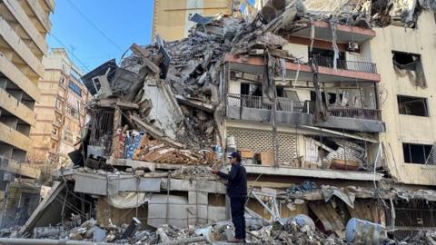 A man stands in front of a building shattered by an airstrike.