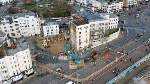 An aerial view of what is left of the Royal Albion Hotel. There is scaffolding around the cream building and three cranes on the road.