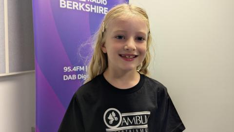 A young girl with light blonde hair and wearing a black T-shirt with white branding. She's standing in front of a sign with the purple BBC Berkshire branding on it.