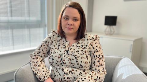 Nikella has shoulder length brown hair. She is sitting on a greychair in a cream-coloured living room. She is wearing a white top with a black and brown spot  pattern. A cabinet with a lamp sitting on top are in the background.
