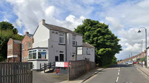 The former Dog and Gunn Inn is a large white building with a brick-built extension behind. It is on a residential road with terraced housing on the other side and a large tree behind it.