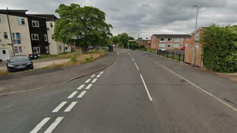 A Google street view image of a residential road. A junction is on the left and behind that is a cream and black coloured block of flats, with a car parked in the car park in front of it and a large tree. On the right are other residential buildings, fences and a bush.