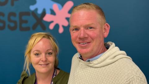 A man and a woman smiling straight at the camera. They are standing in front of a navy wall with the words "POP ESSEX" on it in black writing.