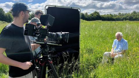 A camera crew are filming Sir David Attenborough sitting in a meadow