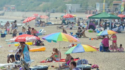 People enjoying the sunshine on the beach in Clacton-on-Sea. There are people sitting beneath colourful umbrellas, lying on towels and walking around the sand.