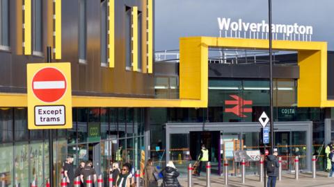 Wolverhampton railway station with yellow hoardings and Wolverhampton in white lettering above. A few people can be seen going into and out of the station.
