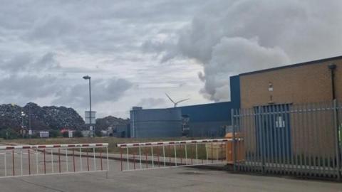 Grey smoke rising from behind a building with large piles of rubbish to the left of it