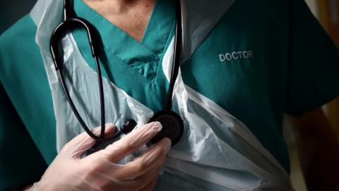 A doctor in green scrubs with a plastic apron over the chest and stethoscope around hi neck.