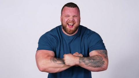 Tom Stoltman, wearing a blue t-shirt and flexing his biceps