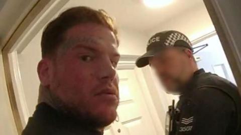 A still taken from bodyworn footage of Codey Nolan's arrest. He is looking angrily at the camera while being led out of a doorway by a police officer wearing a black cap and uniform.