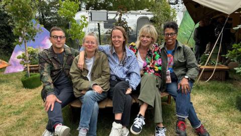 (Left to Right) - Jonathan Rothery, Alison Howe, Emily Eavis, Zoe Ball, Lorna Clarke all sat on a bench backstage at Glastonbury Festival.
