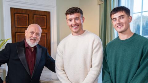 Bill Bailey wearing a suit standing in a room next to two brothers, one wearing a white top and the other wearing a green top