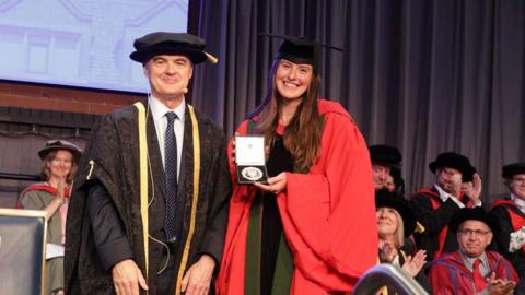 Prof Conchúr Ó Brádaigh is wearing a dark ceremonial gown and hat and Amber Keegan is wearing a bright red graduation gown and mortarboard while holding an open presentation box containing a medal. Several other people in academic dress are seated in the background, some applauding. Dark curtains frame the stage, and a projection screen is visible in the upper left corner.