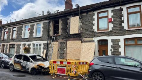 A stone terraced house with fire damage around the windows and doors, and wooden boards covering them. In front are two cars parked, one is badly fire damaged.
