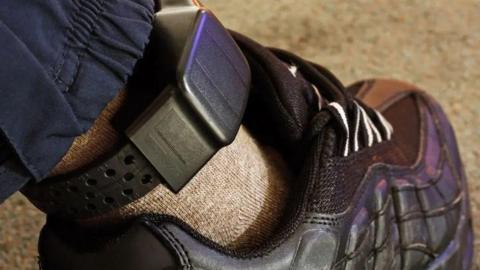 A close up image of a black ankle monitor. It is over a light brown sock on someone's leg above a black shoe