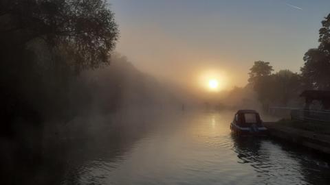 A small boat is moored up on the right-hand side of the picture but the majority of the shot is the river with a thick layer of mist lying over it. The sun is also shrouded in mist in the background and there are trees silhouetted on both sides of the river